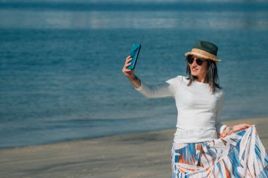 Genç bir kadın sahilde canlı video ya da cep telefonuyla selfie çekiyor.