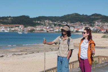 Sırt çantalı çift sahilde yürüyor ve eğleniyor.