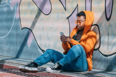 şehirli afro kadın cep telefonuyla grafiti duvarında oturuyor