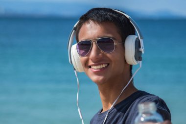 Plajda kulaklıkları ve bir şişe suyu olan rahat genç bir adam.