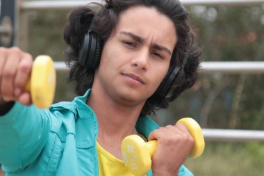 Kulaklıklı genç adam dışarıda halterlerle egzersiz yapıyor.