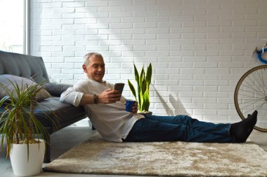 Olgun bir adam evde kahve ve cep telefonuyla kanepede dinleniyor.