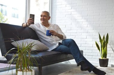 Olgun bir adam evinde kahve ve cep telefonuyla kanepede dinleniyor.