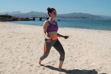 Sahilde dambıllarla egzersiz yapan genç bir kadın.