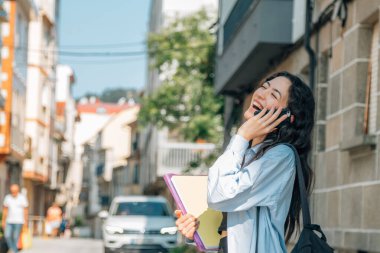 Kitaplı ve sırt çantalı bir öğrenci sokakta telefonla konuşuyor.