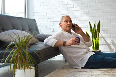 Olgun bir adam evde kahve ve cep telefonuyla kanepede dinleniyor.