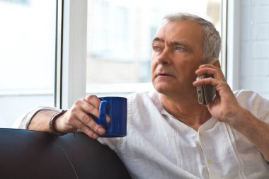 Olgun bir adam evinde kahve ve cep telefonuyla kanepede dinleniyor.