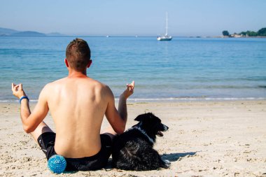 Genç bir adam köpeğiyle plajda meditasyon ya da yoga yapıyor.