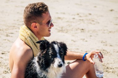 Genç adam sahilde köpeğiyle rahatlıyor ve bir şişe su içiyor.