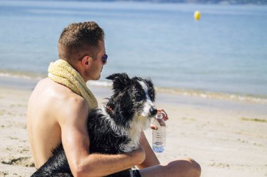 Genç adam sahilde köpeğiyle rahatlıyor ve bir şişe su içiyor.