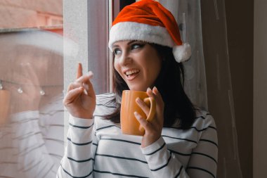 Noel Baba şapkalı genç bir kadın Noel 'de pencerede durup el sallıyor.