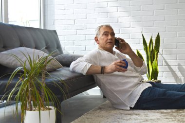 Olgun bir adam evde kahve ve cep telefonuyla kanepede dinleniyor.