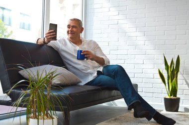 Olgun bir adam evinde kahve ve cep telefonuyla kanepede dinleniyor.