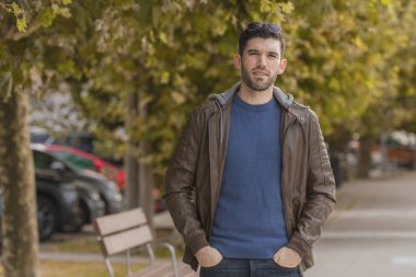 Deri ceketli genç adam sonbaharda parkta yürüyor.