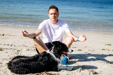 Genç bir adam köpeğiyle plajda meditasyon ya da yoga yapıyor.