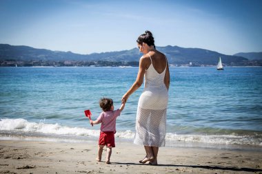 Anne ve oğlu el ele kumsalda yürüyor.