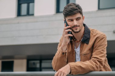 Gülümseyen genç adam sokakta telefonla konuşuyor.