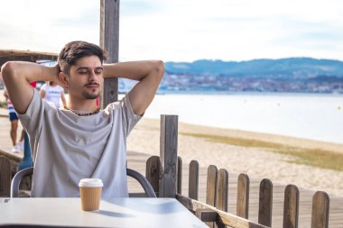 Plaj barındaki genç adam kahve içiyor, dinleniyor ve denize bakıyor.