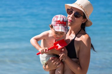 Anne ve küçük çocuğu kumsalda oynuyorlar ve birlikte eğleniyorlar. Dışarıda mutlu bir aile. Anne ve bebek yazın doğada. Deniz kıyısında aile tatili.