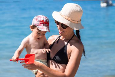 Anne ve küçük çocuğu kumsalda oynuyorlar ve birlikte eğleniyorlar. Dışarıda mutlu bir aile. Anne ve bebek yazın doğada. Deniz kıyısında aile tatili.