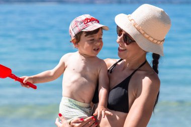 Anne ve küçük çocuğu kumsalda oynuyorlar ve birlikte eğleniyorlar. Dışarıda mutlu bir aile. Anne ve bebek yazın doğada. Deniz kıyısında aile tatili.