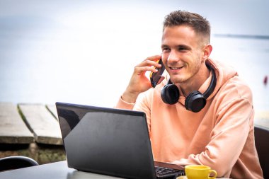 Genç adam dizüstü bilgisayar kullanıyor ve dışarıda telefonla konuşuyor.