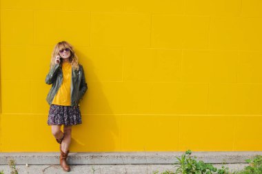 Sarı duvara yaslanmış, cep telefonu olan olgun bir kadın.