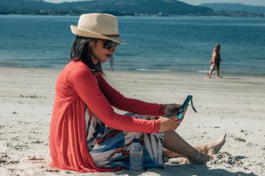 Kumsalda oturmuş akıllı telefonuyla gülümseyen genç kadın.