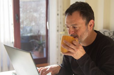 Olgun bir adam iş yerinde bir fincan kahve içerken dizüstü bilgisayarla çalışıyor.