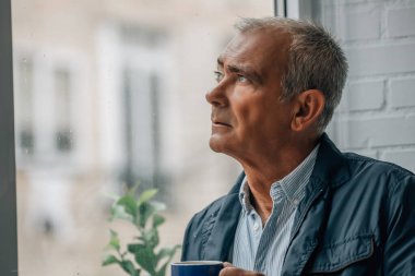 Olgun bir adam elinde kahveyle pencereden dışarı bakıyor.