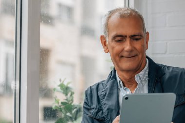 Evde veya iş yerinde dijital tablet kullanan gülümseyen olgun bir adam.