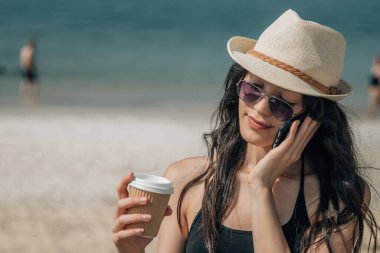 Genç bir kadın sahilde kahve ve cep telefonuyla eğleniyor.