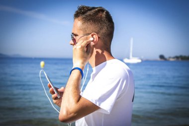 Tatillerde ya da tatillerde kulaklık takan ve telefonla müzik dinleyen genç bir adam.