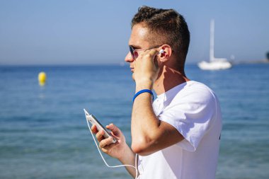 Tatillerde ya da tatillerde kulaklık takan ve telefonla müzik dinleyen genç bir adam.