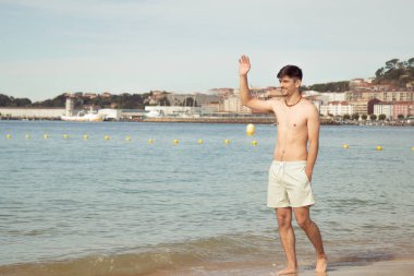 Genç adam kumsalda el sallarken deniz kıyısında yürüyor.