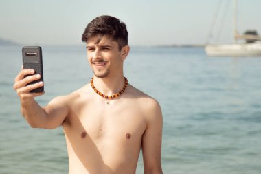 Genç adam sahilde selfie çekiyor ya da cep telefonuyla canlı video çekiyor.