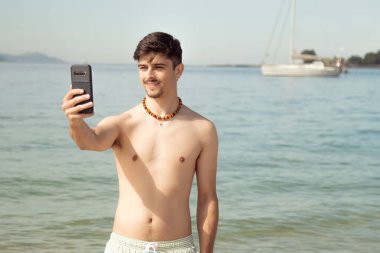 Genç adam sahilde selfie çekiyor ya da cep telefonuyla canlı video çekiyor.