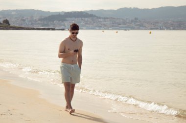 Genç adam plajda yürürken telefonuna bakıyor.