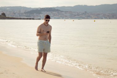 Genç adam plajda yürürken telefonuna bakıyor.