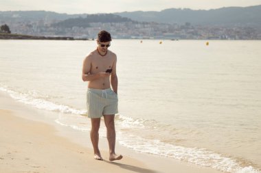Genç adam plajda yürürken telefonuna bakıyor.