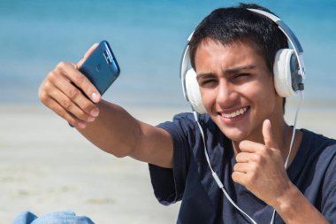 Kulaklıklı ve telefonlu genç adam tamam işareti yaparken selfie çekiyor.