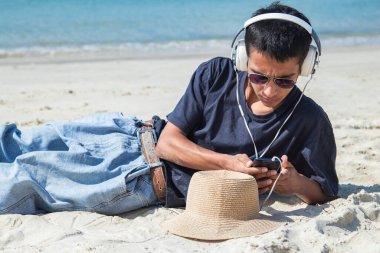 Kulaklığı ve telefonu olan genç bir adam kumsalda kumsalda yatıyor.