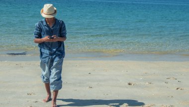 Genç adam plajda yürürken telefonuna bakıyor.