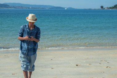 Genç adam plajda yürürken telefonuna bakıyor.