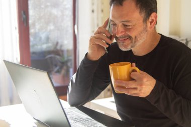 Ofiste ya da iş yerinde bir fincan kahve içerken laptop ve cep telefonuyla çalışan olgun bir adam.