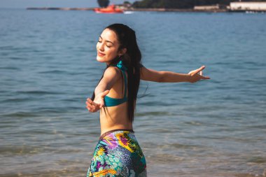 Kollarını kaldırmış gülümseyen genç kadın plajda keyfine bakıyor.