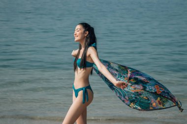 Eşarplı güzel bir kadın kumsalda yürürken rüzgarda sallanıyor.