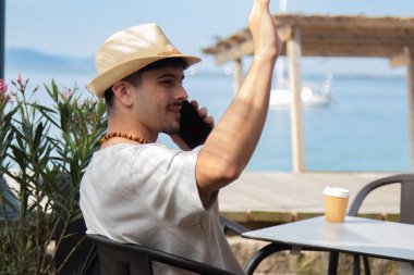 Plaj barında cep telefonuyla konuşan ve el sallayan rahat genç adam.