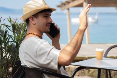 Plaj barında cep telefonuyla konuşan ve el sallayan rahat genç adam.