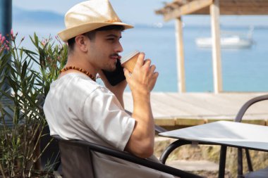 Cep telefonlu rahat genç adam plaj barında kahve içiyor.